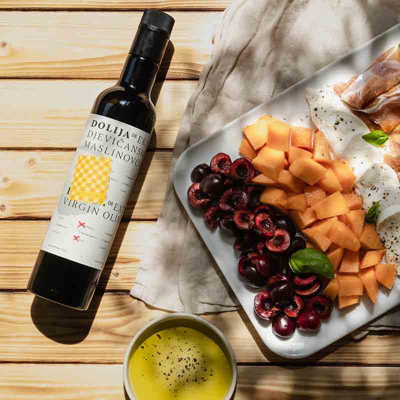 Olivenöl, Früchte, Käse, Schinken auf Holzuntergrund, Dolija-Flasche sichtbar.