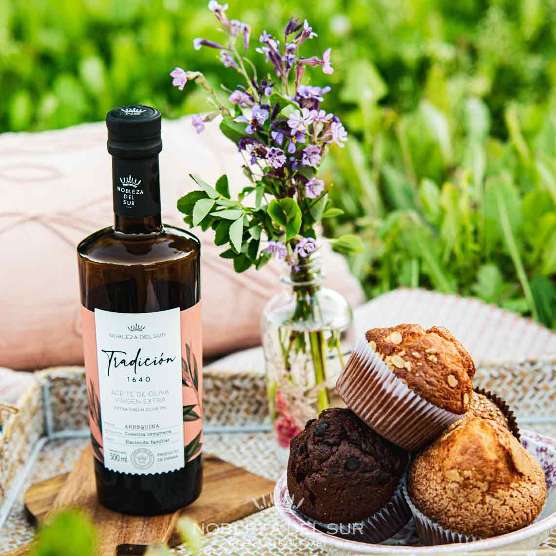 Olivenölflasche, Muffins, Blumenarrangement auf Picknickdecke im Freien.