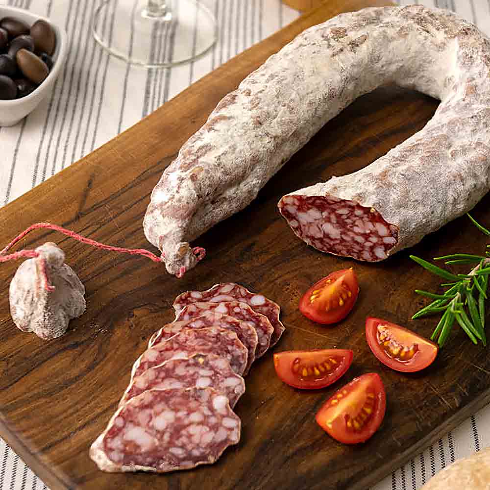 Wurst, Tomaten, Rosmarin auf Holzbrett, Oliven im Hintergrund.
