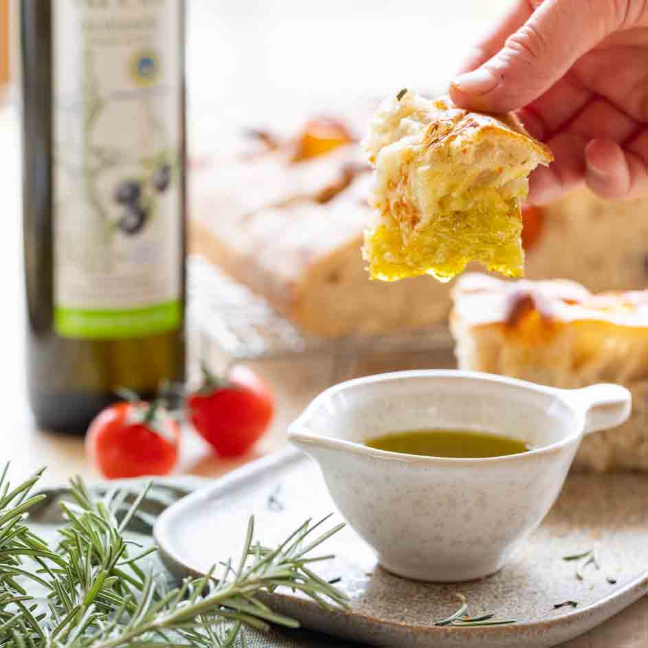Hand taucht Brot in Olivenöl neben Flasche und Tomaten.