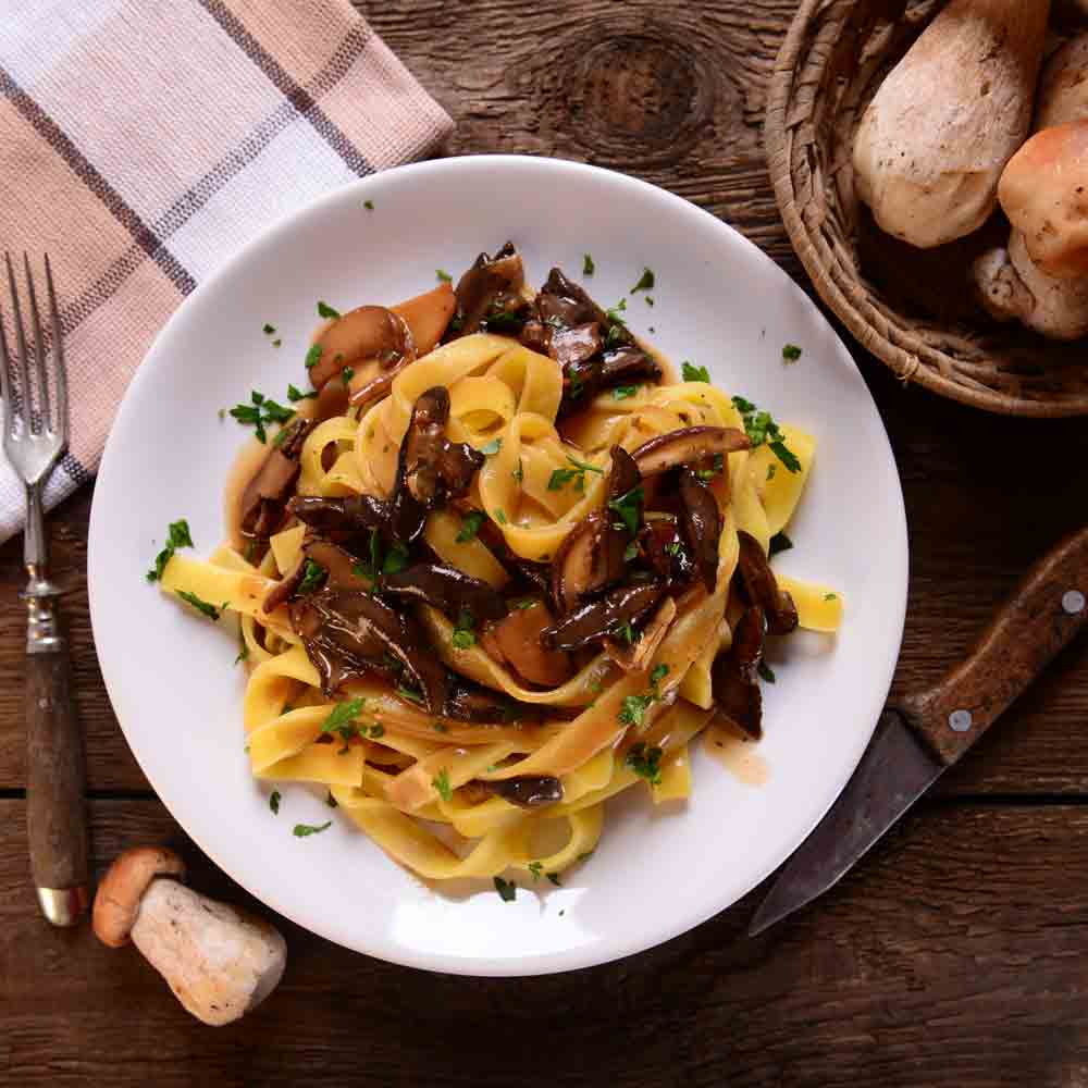 Tagliatelle mit Steinpilzen auf weißem Teller, rustikaler Hintergrund.