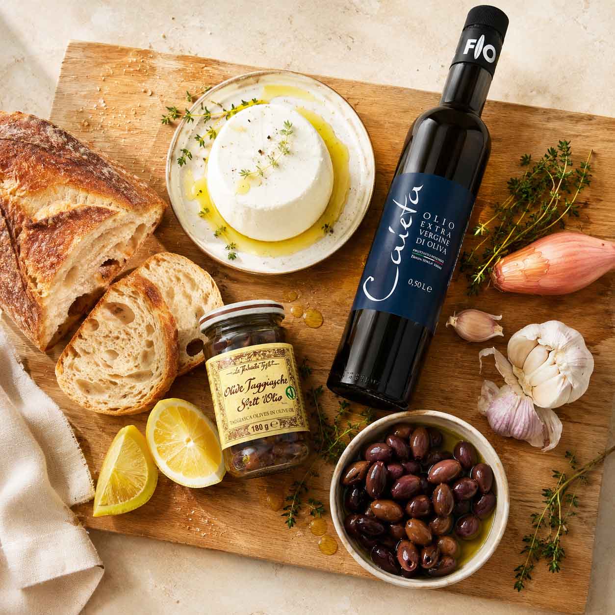 Benötigte Zutaten für das Confit auf einem Schneidebrett, Olivenöl, Taggiasca Oliven, Zitrone, Knoblauch, Schalotte, Thymian, Honig, Ricotta und Frischkäse